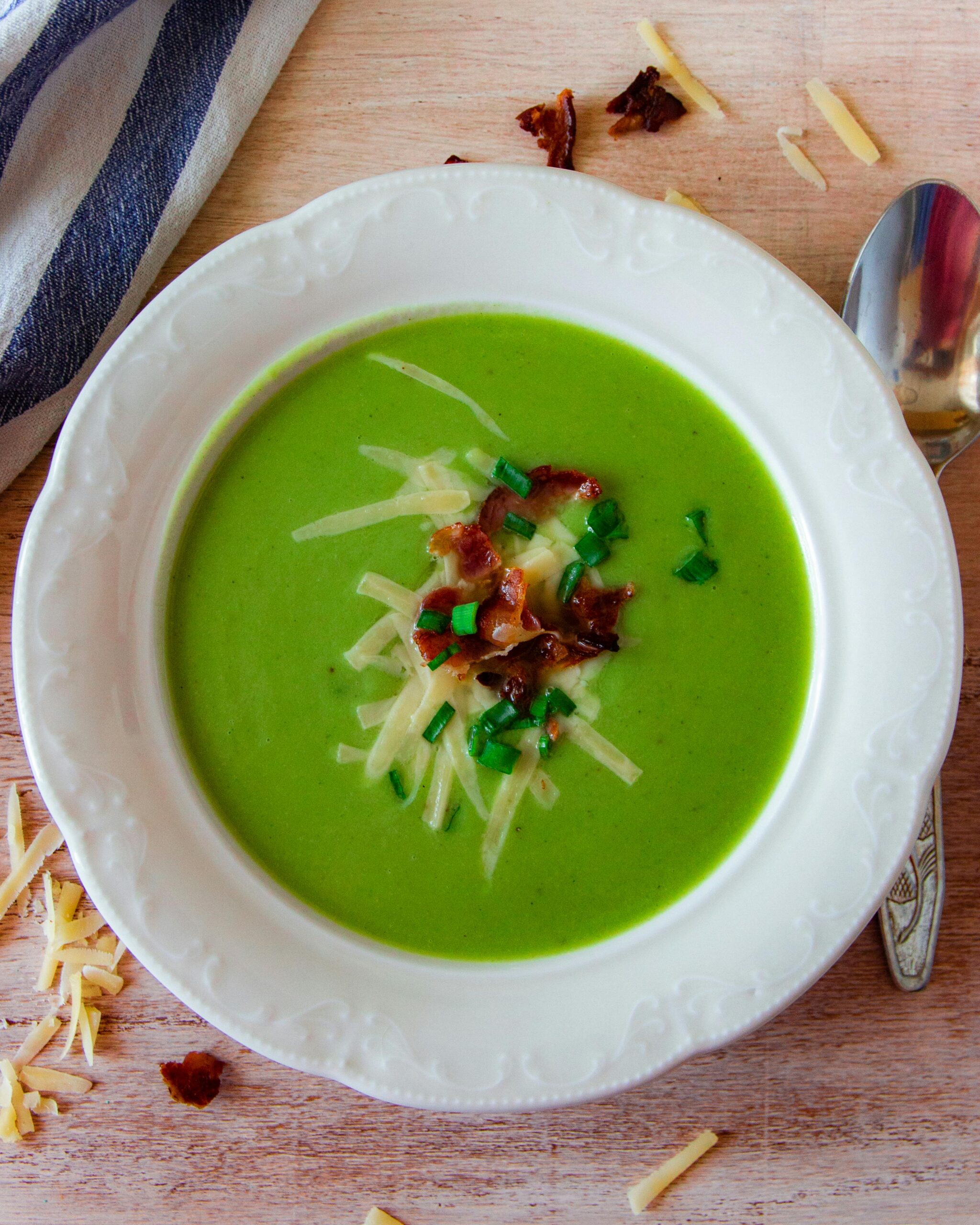 A vibrant green soup garnished with cheese and bacon in a white ceramic bowl on a wooden table.